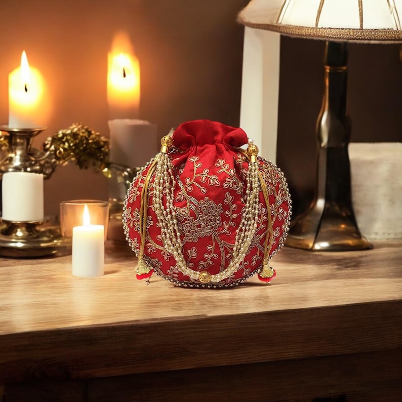 May include: A red, round, embroidered clutch bag with gold and white embellishments. The bag has a drawstring closure and a beaded tassel.