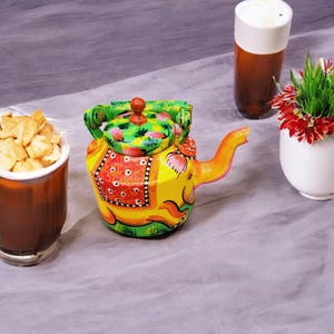 May include: A colorful hand-painted teapot shaped like an elephant. The teapot is yellow, green, red, and orange with black accents. The teapot has a spout and a handle.