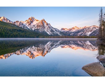 Op de afbeelding: Een vredig landschap met besneeuwde bergen die weerspiegeld worden in een kalm meer. De toppen worden verlicht door de zon, met een heldere blauwe lucht erboven. Het wateroppervlak is glad en creëert een spiegelachtige reflectie.