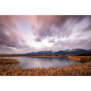 May include: A still lake with a mountain range in the background. The sky is a soft purple with white clouds. The lake is surrounded by tall, brown reeds.