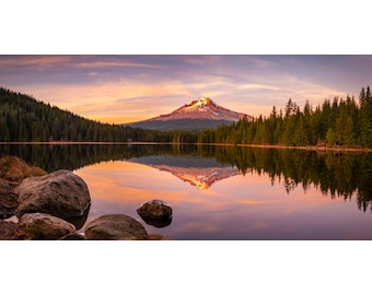 Pode incluir: Uma vista panorâmica de uma montanha coberta de neve refletida em um lago tranquilo ao pôr do sol. A montanha é cercada por uma floresta de árvores de folha perene. O céu é uma mistura vibrante de rosa, roxo e laranja.