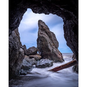 May include: A view of the ocean through a rock formation. The water is a blurry white, and a large rock formation is visible in the distance. The sky is a light blue with some clouds.