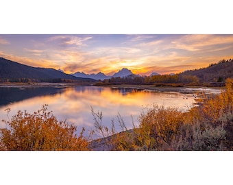 Pode incluir: Uma vista panorâmica de um lago com montanhas ao fundo. O céu é de um laranja e rosa vibrantes, refletindo na água. Em primeiro plano, arbustos e árvores de cor castanho dourado.