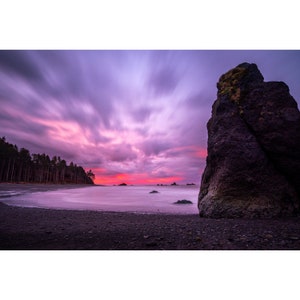 May include: A large, dark rock formation stands on a black sand beach with a purple and pink sky in the background. The water is calm and reflects the colors of the sky.