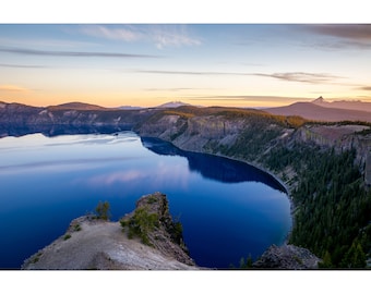 May include: A panoramic view of a deep blue lake surrounded by a rocky, forested shoreline. The lake reflects the sky, creating a mirror-like surface. The distant mountains are visible in the background.