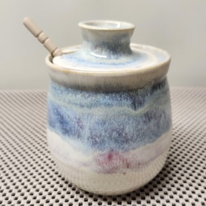 Peut inclure: Un pot en céramique avec un couvercle et une petite cuillère. Le pot présente un dégradé de couleurs bleu, blanc et violet. Le pot est rond avec une petite ouverture en haut. La cuillère est marron clair et repose sur le couvercle.