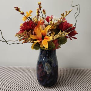 Puede incluir: Un jarrón de cerámica con un esmalte azul oscuro contiene un vibrante arreglo de flores artificiales. El ramo presenta girasoles naranjas, dalias rojas y bayas amarillas, con acentos dorados y verdes. El jarrón mide unos 25 cm de alto.