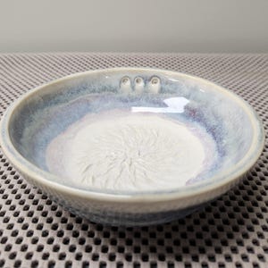 Puede incluir: Un cuenco de cerámica hecho a mano con un exterior texturizado y un interior blanco arremolinado. El borde del cuenco presenta un degradado de tonos azules y morados. Tres pequeñas hendiduras son visibles en el borde. Este cuenco decorativo es adecuado para servir alimentos.