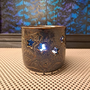Puede incluir: Un portavelas de cerámica marrón oscuro con recortes en forma de estrella. El soporte tiene una superficie texturizada con patrones en relieve. El interior brilla con una luz azul, creando un ambiente cálido. Es un artículo decorativo.