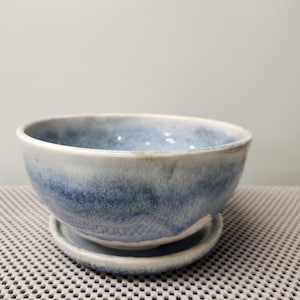 Könnte beinhalten: Ein Keramikschalen- und Untertassen-Set. Die Schale hat eine abgerundete Form mit einer hellblauen und weißen Glasur. Die Untertasse ist eine kleinere, flache Scheibe, ebenfalls mit einer blau-weißen Glasur. Das Set eignet sich zum Servieren von Speisen oder als Dekorationsartikel.