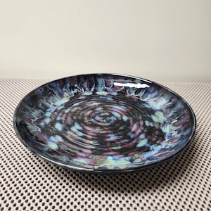 May include: A round ceramic plate featuring a glossy finish with a dark base color and swirling patterns of purple, blue, and light green. The surface has a distinct concentric rippled texture that radiates from the center toward the raised rim.