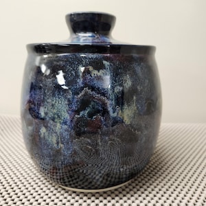 Puede incluir: Una taza de cerámica blanca con un asa negra y el texto "But First, Coffee" en negrita negra. La taza se encuentra sobre un fondo neutro, resaltando su diseño simple.