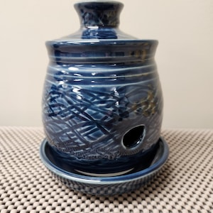 Op de afbeelding: Een donkerblauwe keramische knoflookhouder met een deksel, een kleine opening aan de zijkant en een bijpassende basis. De houder heeft een textuur, geweven patroon. Het keramische stuk staat op een patroon oppervlak.