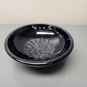 Può includere: Una piccola ciotola rotonda in ceramica blu scuro con un motivo floreale strutturato al centro. La ciotola ha una finitura lucida e tre piccoli fori distanziati vicino al bordo.