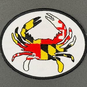 May include: An oval-shaped embroidered patch featuring a crab design. The crab is colored with the red, white, black, and yellow pattern of the Maryland state flag. The patch has a white background and a thick black stitched border edge.