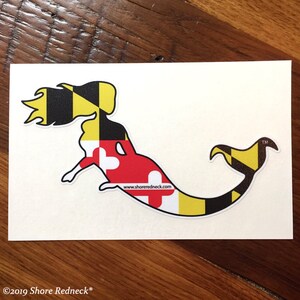 May include: A white sticker with a black and yellow mermaid silhouette. The mermaid is outlined in black and filled with the colors of the Maryland state flag. The text "www.shoredredneck.com" is printed on the sticker. The sticker has a trademark symbol and the text "TM" in the upper right corner.