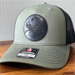 May include: Olive green Richardson 112 trucker hat with a black mesh back. The hat features a circular black patch with a crab design. The hat has a curved brim and an adjustable strap. The Richardson label is visible.