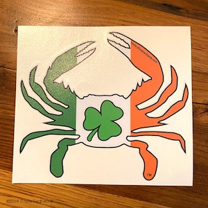 May include: A white sticker with a green and orange crab design. The crab is outlined in black and has a shamrock in the center. The text "www.shoredredneck.com" is printed on the sticker. The sticker also has a trademark symbol and the text "©2019 Shore Redneck".