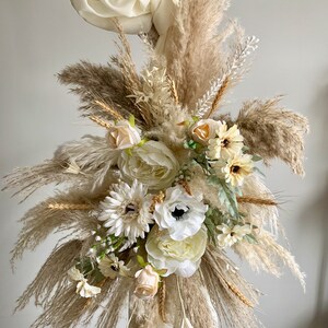 May include: A bouquet of dried pampas grass and white flowers, including roses, anemones, and daisies, arranged in a decorative way. The bouquet is tied with a white ribbon and is displayed against a white background.