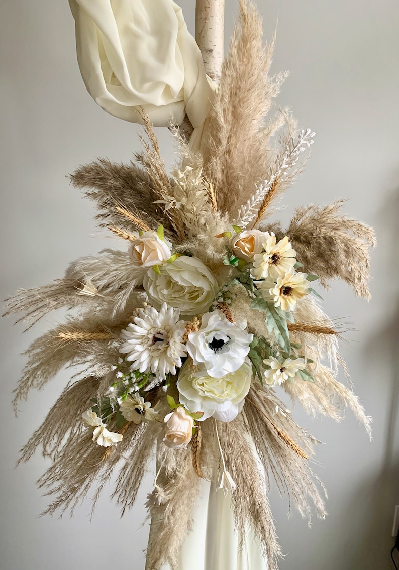 May include: A bouquet of dried pampas grass and white flowers, including roses, anemones, and daisies, arranged in a decorative way. The bouquet is tied with a white ribbon and is displayed against a white background.