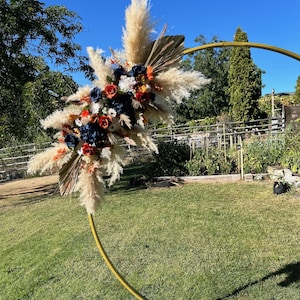 May include: A gold metal arch adorned with a floral display. The arrangement includes pampas grass, navy, orange, and white flowers. The arch is positioned on a grassy lawn against a clear blue sky.