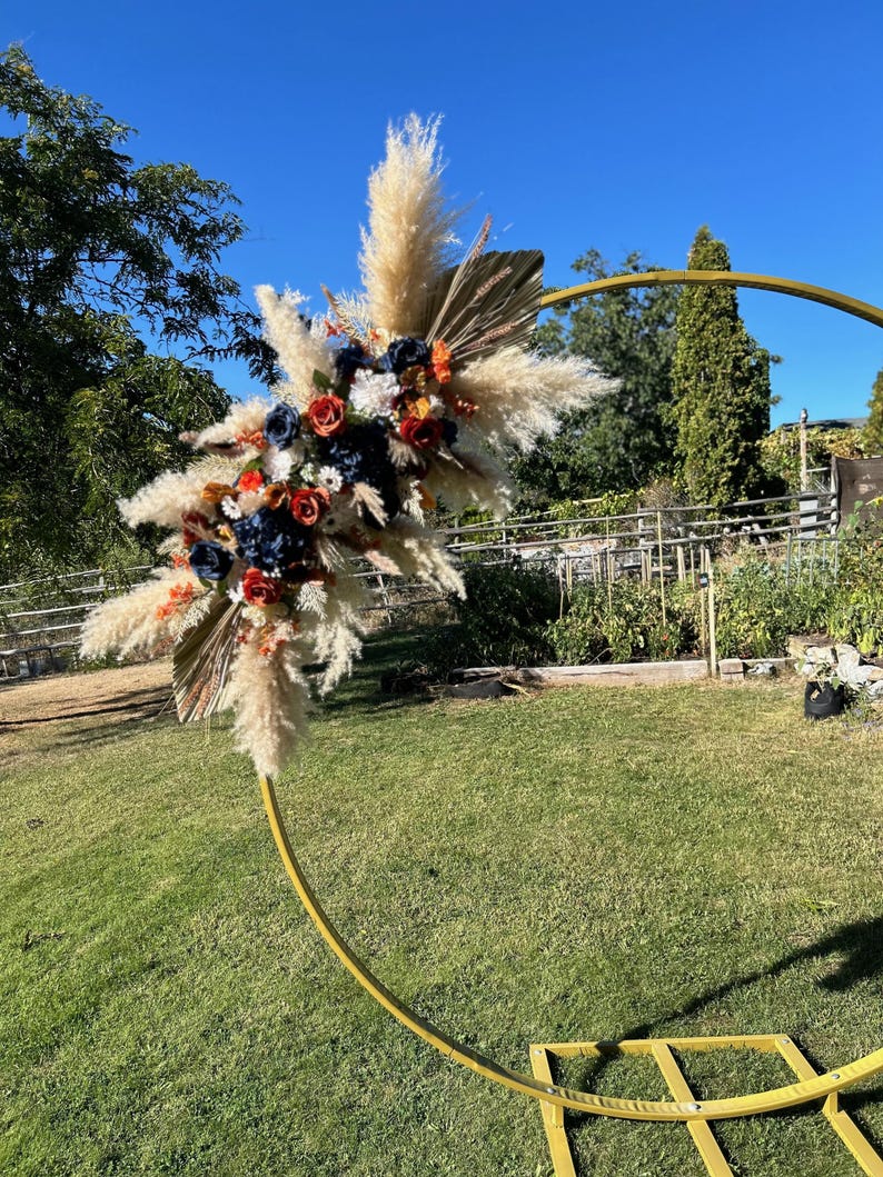 May include: A gold metal arch adorned with a floral display. The arrangement includes pampas grass, navy, orange, and white flowers. The arch is positioned on a grassy lawn against a clear blue sky.