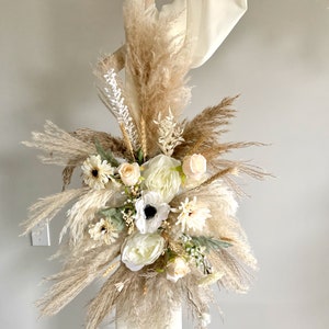 May include: A bouquet of dried pampas grass and white flowers, including anemones, roses, and daisies, arranged in a cluster. The bouquet is tied with a white ribbon and is displayed against a white wall.