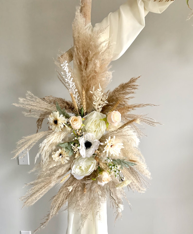 May include: A bouquet of dried pampas grass and white flowers, including anemones, roses, and daisies, arranged in a cluster. The bouquet is tied with a white ribbon and is displayed against a white wall.