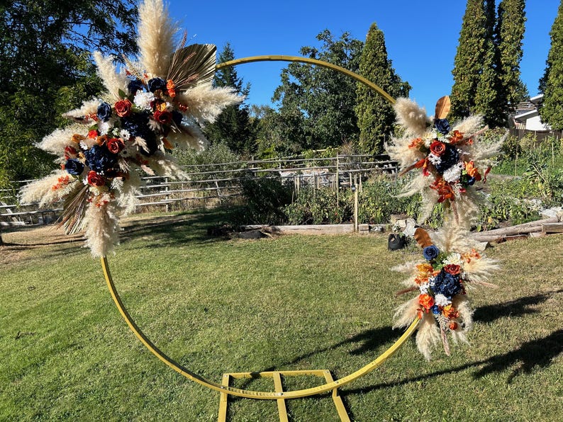 May include: A golden metal wedding arch adorned with pampas grass, navy, red, and orange flowers. The arch stands on a green lawn with trees and a blue sky. The arch is approximately 2.4 meters tall.