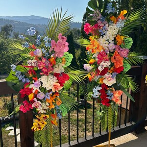 以下が含まれることがあります： 2つの高い金色の金属製のスタンドに、熱帯の花の飾り付けが施されています。飾り付けには、ピンク、オレンジ、イエロー、レッド、ブルーなど、さまざまな色の花が使われています。スタンドは、背景に山脈が見える木製のデッキの上に置かれています。