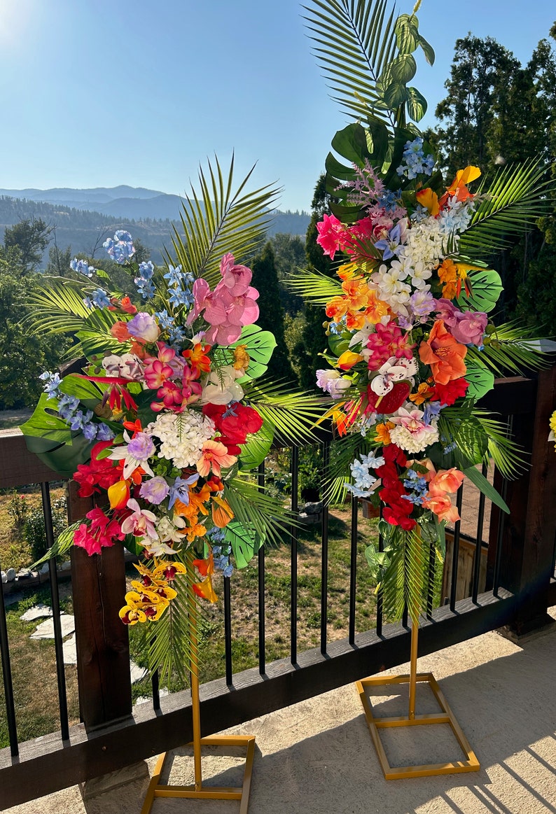 以下が含まれることがあります： 2つの高い金色の金属製のスタンドに、熱帯の花の飾り付けが施されています。飾り付けには、ピンク、オレンジ、イエロー、レッド、ブルーなど、さまざまな色の花が使われています。スタンドは、背景に山脈が見える木製のデッキの上に置かれています。