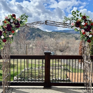May include: A rustic wooden wedding arch decorated with burgundy, white, and peach flowers, greenery, and vines. The arch is set up outdoors on a wooden deck with a view of a mountain range in the background.