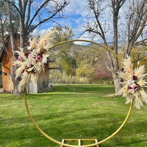 May include: A gold metal archway decorated with dried pampas grass and pink and burgundy flowers. The archway is set up outdoors in a grassy area with trees and a wooden shed in the background.