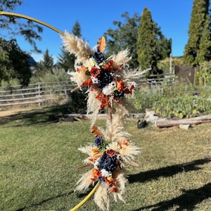 May include: A golden arch adorned with floral arrangements. The arrangements include pampas grass, navy blue, orange, and red flowers. A wooden fence and trees are visible in the background, under a clear blue sky.