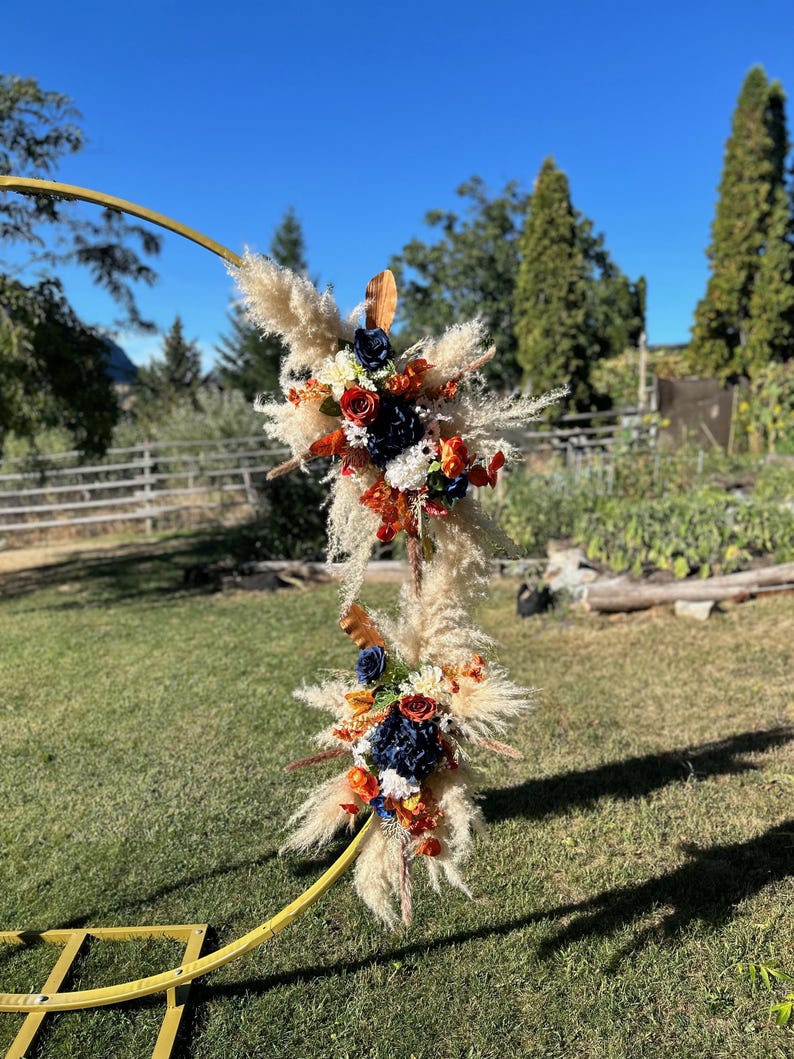 May include: A golden arch adorned with floral arrangements. The arrangements include pampas grass, navy blue, orange, and red flowers. A wooden fence and trees are visible in the background, under a clear blue sky.