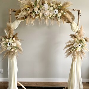 May include: A rustic wooden archway decorated with dried pampas grass, white flowers, and greenery. The archway is adorned with white fabric drapes on each side.