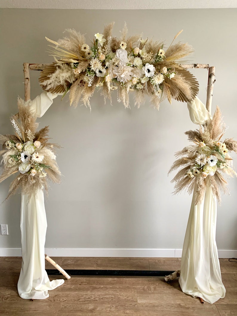 May include: A rustic wooden archway decorated with dried pampas grass, white flowers, and greenery. The archway is adorned with white fabric drapes on each side.
