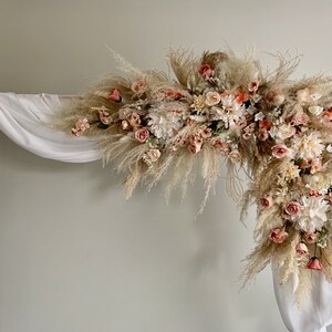 May include: A decorative archway adorned with a lush arrangement of dried pampas grass, peach-colored roses, and white flowers. The arch is draped with white fabric, creating a soft, romantic aesthetic, perfect for weddings or special events.