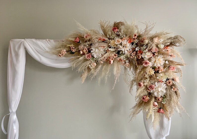 May include: A decorative archway adorned with a lush arrangement of dried pampas grass, peach-colored roses, and white flowers. The arch is draped with white fabric, creating a soft, romantic aesthetic, perfect for weddings or special events.