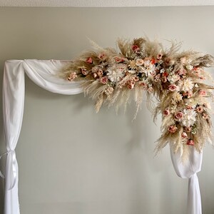 May include: A white wedding arch decorated with a floral arrangement of pampas grass, roses, and other flowers in shades of pink, peach, and cream. White fabric drapes from the arch.