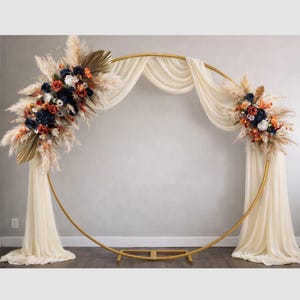 May include: A gold metal wedding arch with a circular design. The arch is decorated with cream-coloured fabric and floral arrangements in shades of navy, orange, and white. Dried pampas grass and palm leaves add texture.
