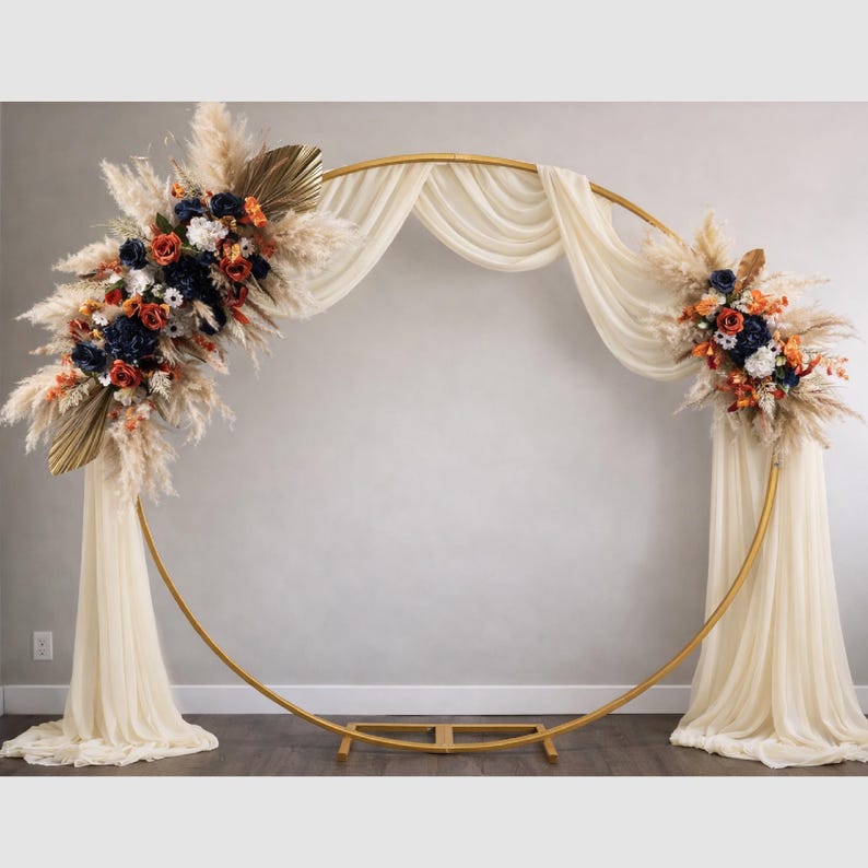 May include: A gold metal wedding arch with a circular design. The arch is decorated with cream-coloured fabric and floral arrangements in shades of navy, orange, and white. Dried pampas grass and palm leaves add texture.