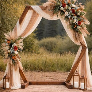 May include: A hexagonal wooden wedding arch draped with sheer beige fabric, adorned with pampas grass, eucalyptus, and red and white roses. Three metal lanterns with white pillar candles sit on a round woven rug at the base of the structure.