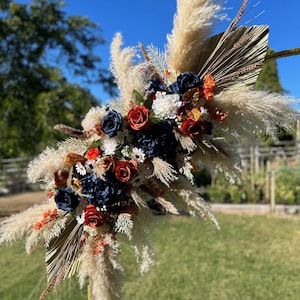 May include: A decorative floral arrangement with navy, red, and white flowers, dried palm leaves, and fluffy pampas grass. The arrangement is set against a blue sky and green grass.