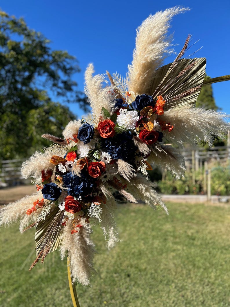 May include: A decorative floral arrangement with navy, red, and white flowers, dried palm leaves, and fluffy pampas grass. The arrangement is set against a blue sky and green grass.