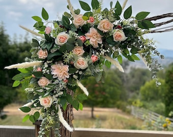 May include: A wedding arch decorated with blush pink roses, white dahlias, and greenery. The arch is made of natural wood branches and is adorned with pampas grass.