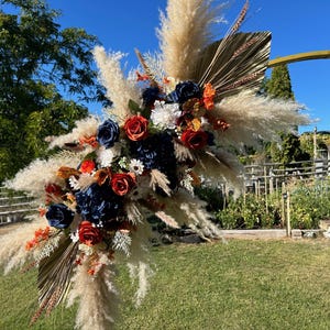 May include: A large floral arrangement with pampas grass, navy blue and red roses, and dried palm leaves. The arrangement is set against a blue sky and green grass, suitable for weddings or special events.