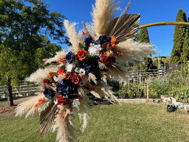 May include: A large floral arrangement with pampas grass, navy blue and red roses, and dried palm leaves. The arrangement is set against a blue sky and green grass, suitable for weddings or special events.