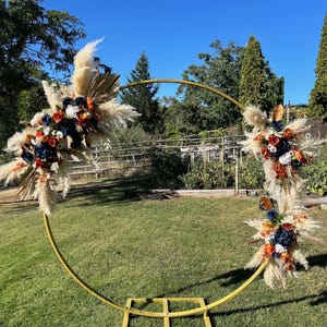 May include: A gold metal wedding arch decorated with floral arrangements. The arch is adorned with pampas grass, dried palm leaves, and colorful flowers in shades of navy, orange, and red. The arch is set on a green lawn under a clear blue sky.