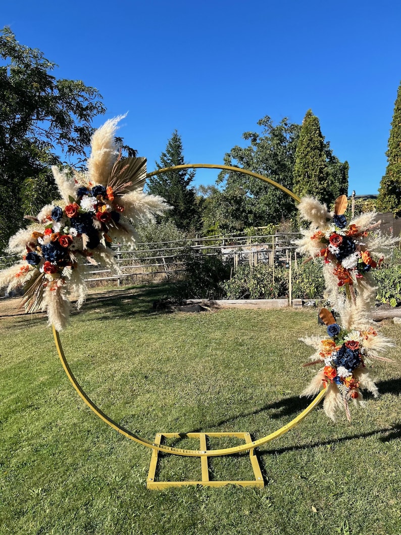 May include: A gold metal wedding arch decorated with floral arrangements. The arch is adorned with pampas grass, dried palm leaves, and colorful flowers in shades of navy, orange, and red. The arch is set on a green lawn under a clear blue sky.